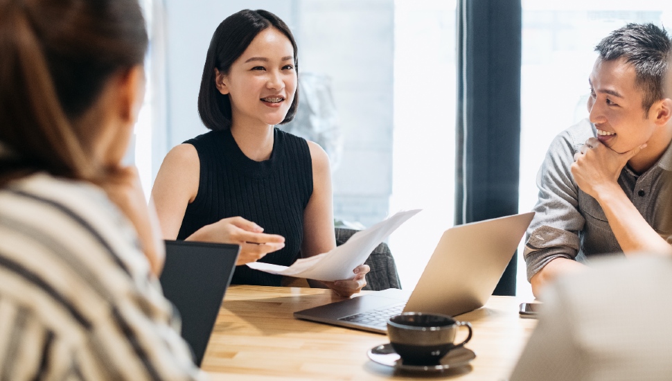 An Asian female adult chatting with two people while seated at the same table.