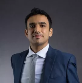 Person in a blue suit and tie smiling against a gray background.