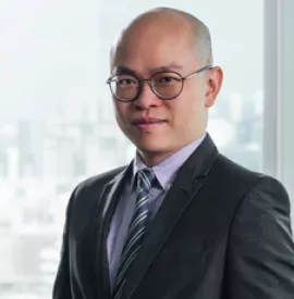 Smiling man in suit with glasses, standing indoors with a city skyline in the background.
