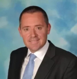 Man in a suit with a light blue tie against a cloudy sky background.