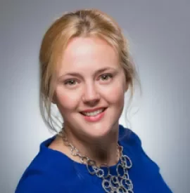 Smiling person with light hair in a blue top against a gray background.