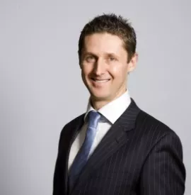 Smiling man in a dark suit and blue tie, standing against a gray background.