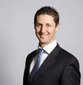 Smiling man in a dark suit and blue tie, standing against a gray background.