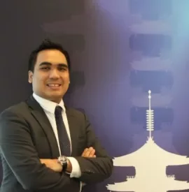 Man in a suit smiling with folded arms, standing in front of a blue backdrop with a pagoda design.