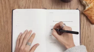 Person writing in a planner next to a croissant on a wooden table.