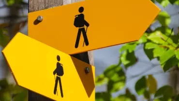 Two yellow hiking trail signs with backpack icons on a wooden post in a forest setting.