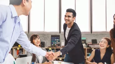 A group of colleagues smiling and shaking hands during a meeting in a bright office.