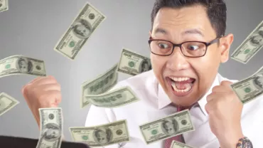 Man celebrating while dollar bills fly around him, looking excited at computer.