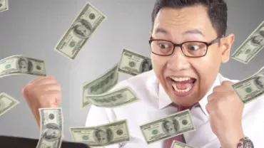 Man celebrating while dollar bills fly around him, looking excited at computer.