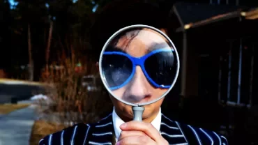 Person in sunglasses holding a magnifying glass to their face outdoors.