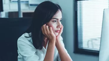 Woman in a white shirt looking at a computer screen, appearing thoughtful.