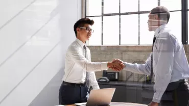 Two men in an office shaking hands, one with glasses and both in business attire.