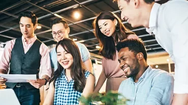 A group of six people smiling and collaborating around a laptop in an office.