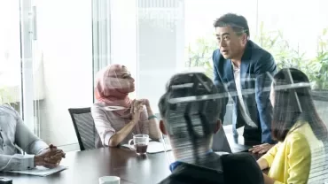 A diverse group of people in a business meeting within a glass-walled office.