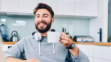 Man sitting in a kitchen, smiling and holding a coffee mug, wearing headphones around his neck.