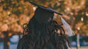 Graduate in cap and gown standing with blurred autumn leaves in the background.