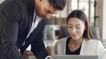 Two colleagues engaged in a discussion over a laptop in an office setting.