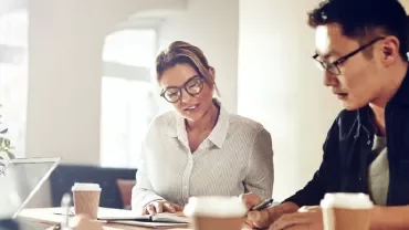 Two people in an office setting, collaborating with laptops and coffee cups on the table.