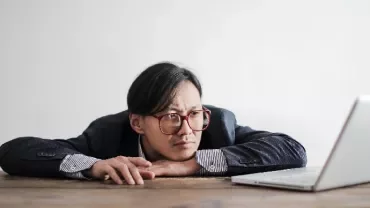Man in glasses staring thoughtfully at a laptop on a wooden table.