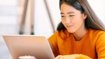 Woman in orange sweater using a laptop, seated indoors.