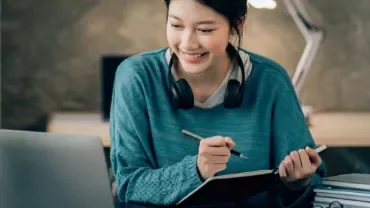 Woman in blue sweater studying with laptop and notebook.