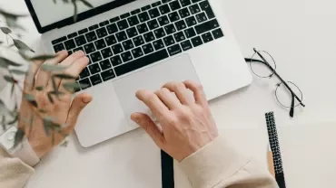 Top view of hands typing on a laptop, with glasses and a notebook on a white desk.