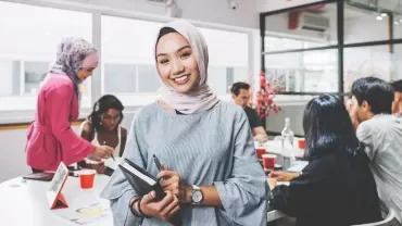 Smiling woman in office attire holding a notebook, with colleagues collaborating in the background.