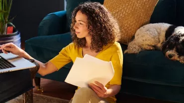 Woman in yellow shirt working on laptop with papers, dog resting on couch.