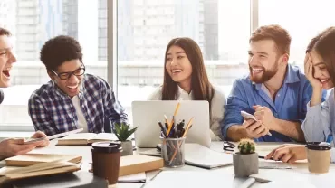 Group of five young adults laughing and talking while working together at a table, with a laptop and coffee cups.