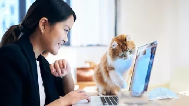 Woman smiling at laptop with curious cat beside her.