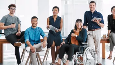 A diverse group of colleagues in an office, smiling and holding devices, with one person in a wheelchair.