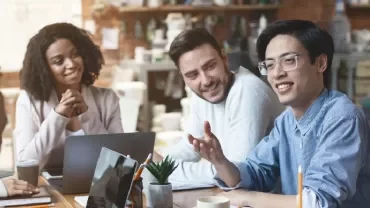 Group of people smiling and discussing at a meeting table with laptops and coffee cups.