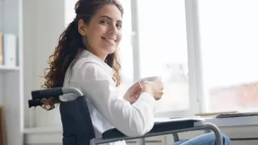 Woman in wheelchair smiling while holding a coffee cup at her desk.
