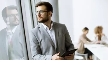 Man in a suit holding a tablet, standing by a window, with colleagues working in the background.