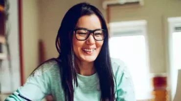 Woman in glasses smiling at a computer screen in a cozy room.