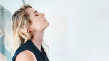 Woman in a blue top leaning back against glass, eyes closed.