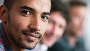 A man with a plaid shirt looking at the camera with two people in the background.