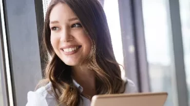 Smiling woman with long hair holding a tablet by a window.