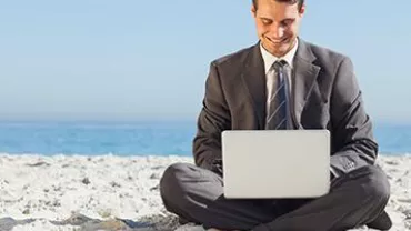 Businessman sitting on beach with laptop, smiling.