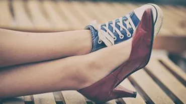 Close-up of legs wearing mismatched shoes: a red high heel and a blue sneaker, resting on a wooden bench.
