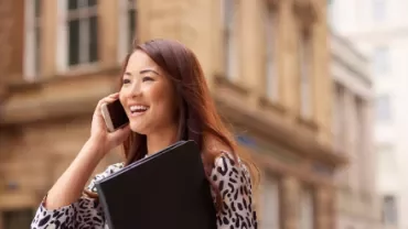 Smiling woman on phone holding a black folder, standing outdoors.