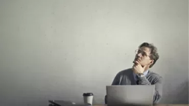 Man sitting at a desk with a laptop, thinking and looking upwards.