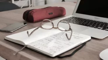 Notebook with eyeglasses on a desk next to a laptop and pencil case.
