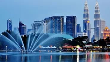 Night view of a city skyline with tall towers and a fountain in the foreground.
