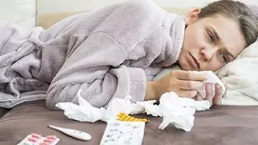 Person lying on a sofa in a robe, surrounded by tissues and medication.