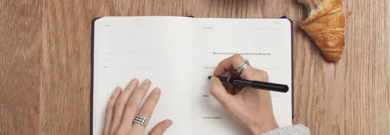 Person writing in a planner next to a croissant on a wooden table.