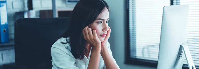 Woman in a white shirt looking at a computer screen, appearing thoughtful.