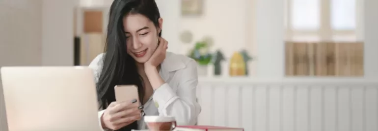 Woman smiling at her phone with a laptop and coffee on the table