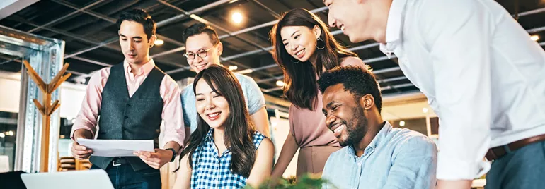 A group of six people smiling and collaborating around a laptop in an office.