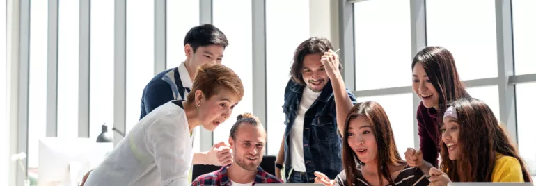 A group of people gathered around a laptop, looking surprised and delighted in a bright office setting.